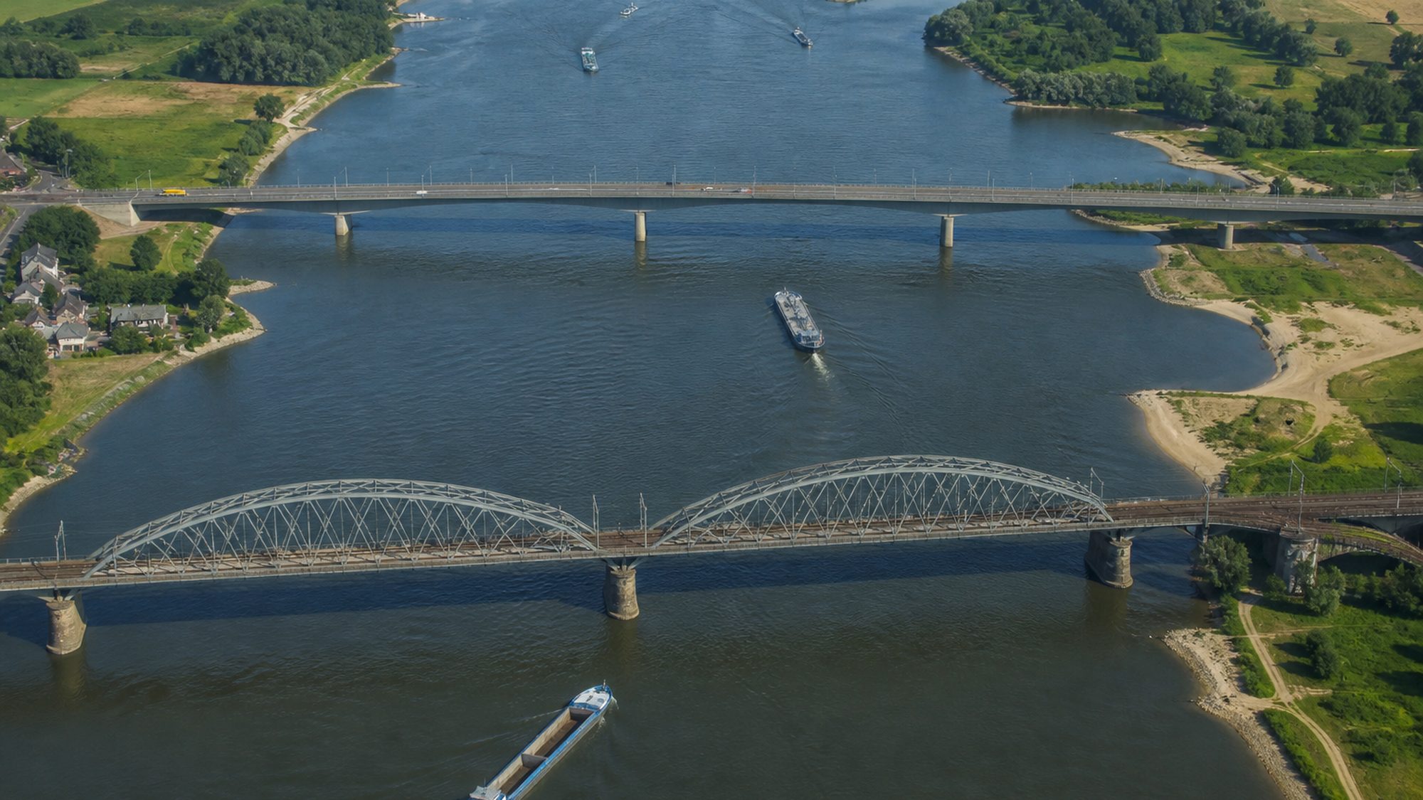 Brede rivier met twee bruggen van bovenaf, vrachtschepen en groene oevers