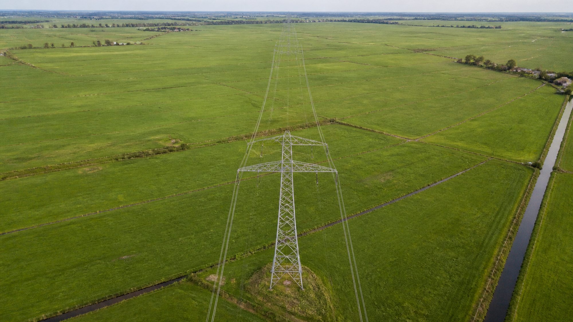 Hoogspanningsmast in vlak weiland, schuin van boven, met leidingen naar de horizon
